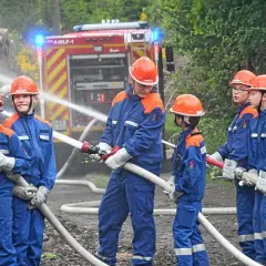 Jugendfeuerwehr übt praktische Ausbildungsideen: Kinder in blauen Uniformen und Helmen halten Wasserschläuche, ein Feuerwehrauto im Hintergrund.