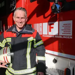 Ein lächelnder hauptamtlicher feuerwehrkommandant in Uniform hält seinen Helm vor einem roten Feuerwehrauto.
