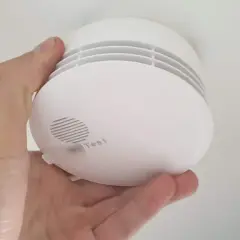 Ein weißer Rauchmelder wird im Keller von einer Hand gehalten. Er hat einen Testknopf und Lüftungsschlitze.