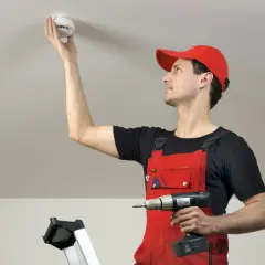 Mann in roter Latzhose montiert Rauchmelder an Decke. Rauchmelder retten Leben – ein wichtiger Schritt für Ihre Sicherheit.