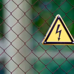 Gefahr! Gelbes Warnschild mit Blitzsymbol auf Drahtzaun. Bei einem leichten Stromschlag was tun? Sofort Abstand halten und Hilfe holen.