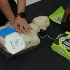 Schulung an einem Trainings-Defibrillator mit Reanimationspuppe. Die Hände üben die Herzdruckmassage. Regelmäßige **Defibrillator Wartung** ist wichtig.