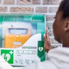 Frau öffnet Defibrillator-Gehäuse, um den Standort zu finden. Gelbes Gerät mit Herzsymbol sichtbar.