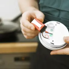 Batteriewechsel am Rauchmelder: Eine Hand hält eine rote 9V-Batterie, bereit zum Einsetzen in das weiße Gerät.