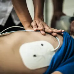 Hände führen eine Herzmassage durch, während ein AED-Einsatz vorbereitet wird. Ein weißes Elektrodenthermometer ist auf der Brust platziert.
