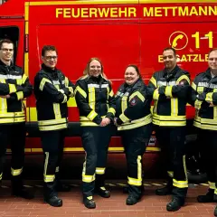 Feuerwehrleute in Mettmann, alle Altersgruppen, stehen stolz vor ihrem roten Einsatzfahrzeug.