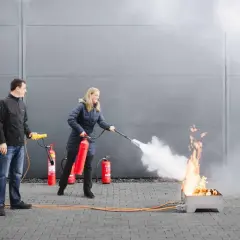 Frau übt mit Feuerlöscher bei Fortbildung Brandschutzhelfer. Mann beobachtet.