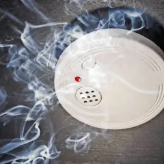 Ein weißer rauchmelder oder feuermelder mit roter LED auf dunklem Grund, umgeben von aufsteigendem Rauch.