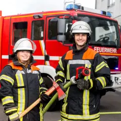 Zwei Feuerwehrleute vor einem roten Feuerwehrauto. Sie tragen Schutzkleidung und Helme. Die Frau hält eine Axt, der Mann einen Schlauch. So wird man Feuerwehrmann oder Feuerwehrfrau.