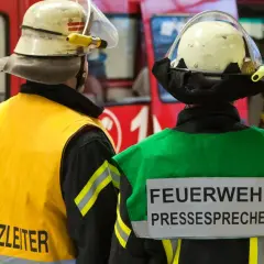 Zwei Feuerwehrleute stehen vor einem roten Fahrzeug. Einer trägt eine gelbe Weste mit der Aufschrift "Einsatzleiter", der andere eine grüne Weste mit der Aufschrift "FEUERWEHR PRESSESPRECHER".