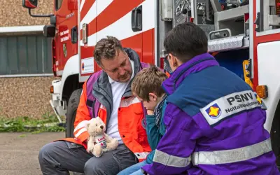 Ein Mann mit einem Teddybär spricht mit einem Kind neben einem Feuerwehrauto. Eine Person mit PSNV-Aufschrift auf der Jacke sitzt daneben.