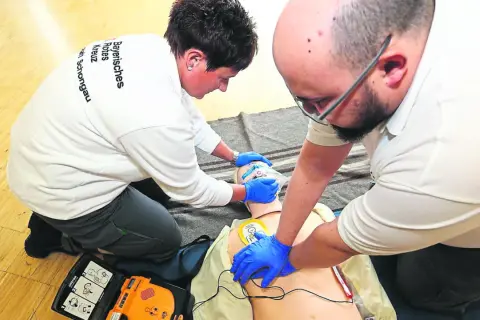 Zwei Personen üben die Defibrillator Anwendung an einer Puppe. Ein Mann führt Herzdruckmassage durch, während eine Frau die Beatmungsmaske hält.