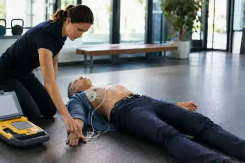 Sanitäterin klebt AED-Elektroden auf Brust eines Mannes, der auf dem Boden liegt. Ein Defibrillator steht daneben.