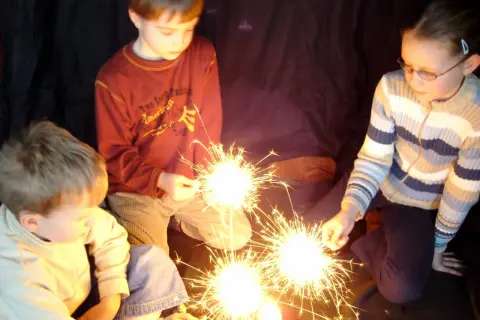 Kinder halten Wunderkerzen. Ein Junge im roten Pullover, ein Mädchen mit Brille und ein kleiner Junge im hellen Pulli genießen den Zauber. Ein schöner Moment, der an die Grundschule erinnert.