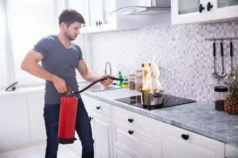 Mann löscht Topfbrand in der Küche mit Feuerlöscher. Dies ist ein Beispiel für Brandklasse F, Fettbrand.