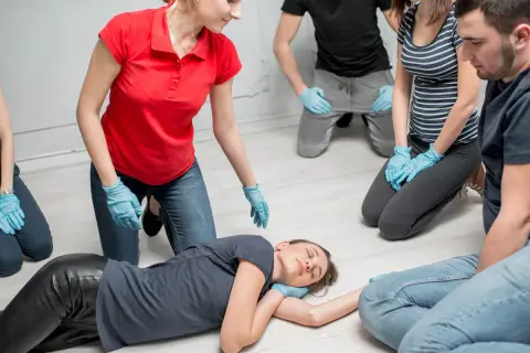 Erste-Hilfe-Kurs: Eine Frau wird in die stabile Seitenlage gebracht. Mehrere Personen mit blauen Handschuhen beobachten aufmerksam.