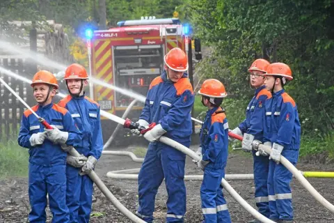 Jugendfeuerwehr übt praktische Ausbildungsideen: Kinder in blauen Uniformen und Helmen halten Wasserschläuche, ein Feuerwehrauto im Hintergrund.