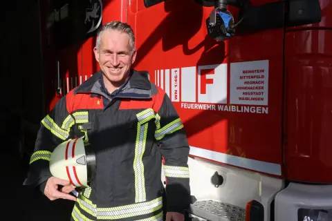 Ein lächelnder hauptamtlicher feuerwehrkommandant in Uniform hält seinen Helm vor einem roten Feuerwehrauto.