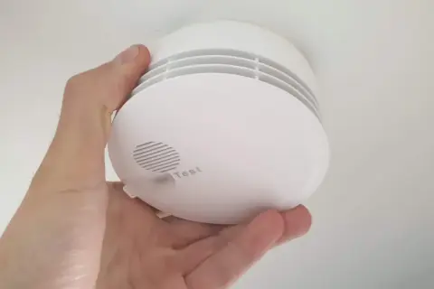 Ein weißer Rauchmelder wird im Keller von einer Hand gehalten. Er hat einen Testknopf und Lüftungsschlitze.