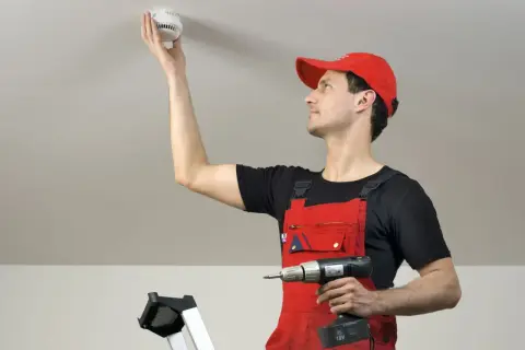 Mann in roter Latzhose montiert Rauchmelder an Decke. Rauchmelder retten Leben – ein wichtiger Schritt für Ihre Sicherheit.