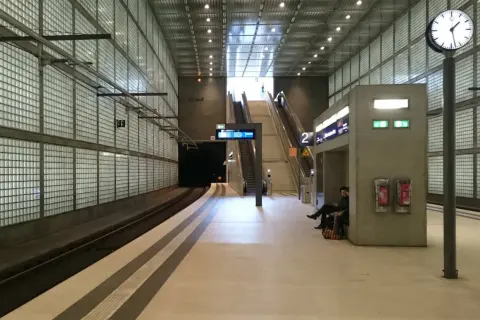 Moderne U-Bahn-Station mit Glaswänden, Rolltreppen und einer Uhr. Die Notbeleuchtung ist gemäß Vorschriften installiert.