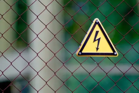 Gefahr! Gelbes Warnschild mit Blitzsymbol auf Drahtzaun. Bei einem leichten Stromschlag was tun? Sofort Abstand halten und Hilfe holen.