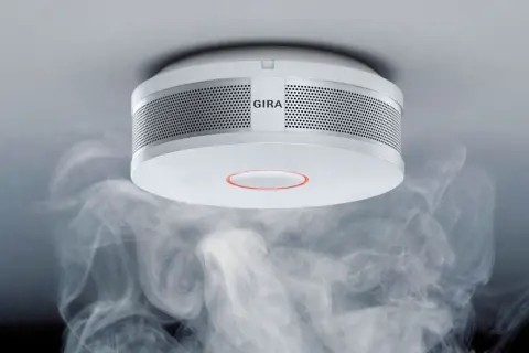 Ein Gira Rauchmelder mit orange blinkendem Ring, umgeben von aufsteigendem Rauch.