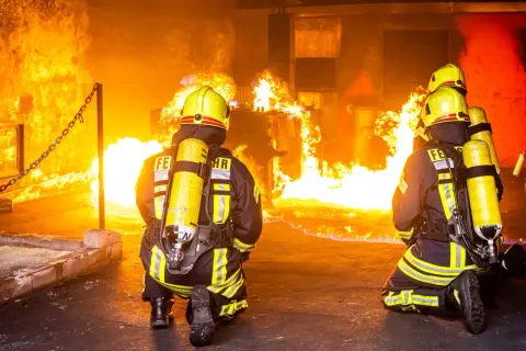 Zwei Feuerwehrleute in voller Montur knien vor einem intensiven Feuer. Ihre Ausbildung bei der Berufsfeuerwehr bereitet sie auf solche Einsätze vor.