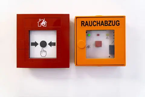 RWA Anlagen Wartung: Rote Brandmeldeanlage und orange Rauchabzugseinheit an weißer Wand.