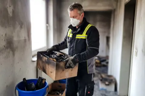 Mann mit Maske sortiert Brandgut in Karton. Wohnung nach Brand unbewohnbar was tun?