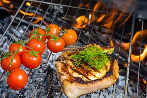 Ein saftiges Kotelett mit Dill und Kirschtomaten auf einem Grill im Haus. Flammen lodern im Hintergrund.