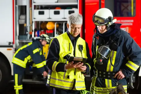 Feuerwehrleute besprechen Einsatzdetails. Ein Mann in Warnweste prüft ein Tablet, während ein Kollege in voller Montur mit Atemschutzmaske daneben steht. UVV Feuerwehr wird beachtet.