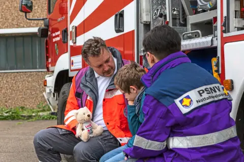 Ein Mann mit einem Teddybär spricht mit einem Kind neben einem Feuerwehrauto. Eine Person mit PSNV-Aufschrift auf der Jacke sitzt daneben.