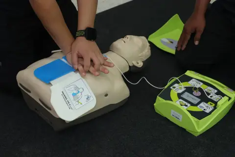 Schulung an einem Trainings-Defibrillator mit Reanimationspuppe. Die Hände üben die Herzdruckmassage. Regelmäßige **Defibrillator Wartung** ist wichtig.