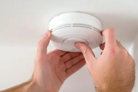 Hände drücken auf den Knopf eines weißen Rauchmelders an der Decke, um den test rauchmelder durchzuführen.