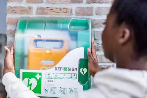 Frau öffnet Defibrillator-Gehäuse, um den Standort zu finden. Gelbes Gerät mit Herzsymbol sichtbar.
