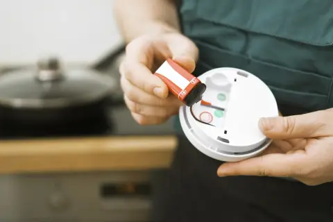 Batteriewechsel am Rauchmelder: Eine Hand hält eine rote 9V-Batterie, bereit zum Einsetzen in das weiße Gerät.