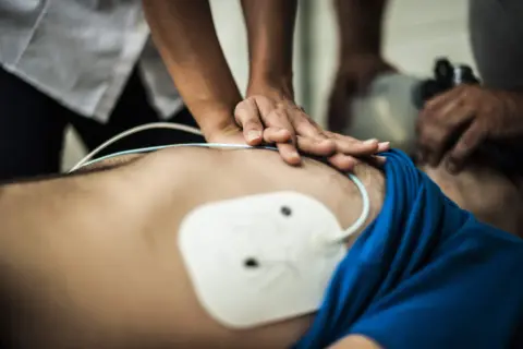 Hände führen eine Herzmassage durch, während ein AED-Einsatz vorbereitet wird. Ein weißes Elektrodenthermometer ist auf der Brust platziert.