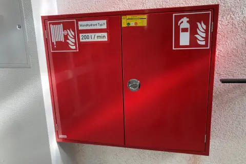 Roter Schrank für Brandschutz in einer Arztpraxis: Wandhydrant Typ F, 200 l/min, und Feuerlöscher-Symbol.