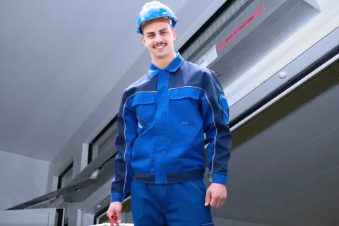 Ein Handwerker in blauer Arbeitskleidung und Helm trägt einen Werkzeugkasten. Er ist bereit für den Einsatz nach DIN 14677.