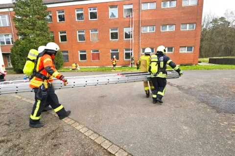 Feuerwehr-Messe: Einsatzkräfte transportieren eine Leiter vor einem Backsteingebäude. Eine Person steigt bereits eine Leiter zum Fenster empor.