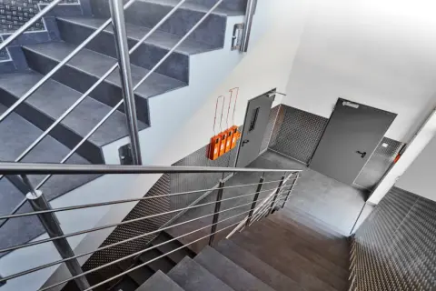 Moderner Treppenaufgang mit Metallgeländern und grauen Stufen. An der Wand hängen orangefarbene Geräte, die Teil des Rauchabzug Treppenhaus Systems sind.
