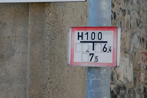 Nahaufnahme eines Hydrantenschilds mit der Aufschrift "H100 T 6,4 7,5". Das Schild ist an einem Metallpfosten befestigt, daneben eine raue Betonwand.