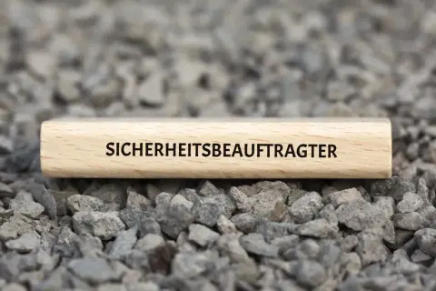 Ein Holzstab mit der Aufschrift "SICHERHEITSBEAUFTRAGTER KITA" liegt auf grauem Schotter.