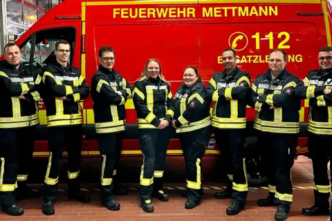 Feuerwehrleute in Mettmann, alle Altersgruppen, stehen stolz vor ihrem roten Einsatzfahrzeug.