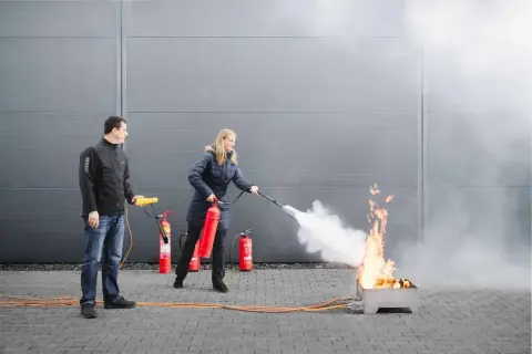 Frau übt mit Feuerlöscher bei Fortbildung Brandschutzhelfer. Mann beobachtet.