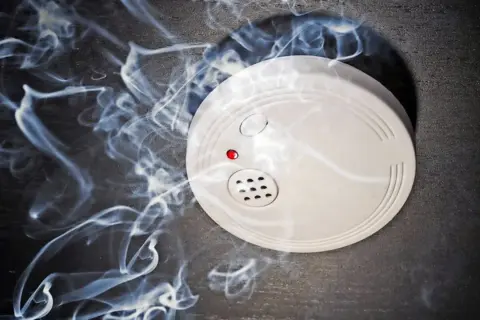 Ein weißer rauchmelder oder feuermelder mit roter LED auf dunklem Grund, umgeben von aufsteigendem Rauch.
