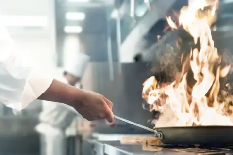 Ein Koch wirbelt eine brennende Pfanne in einer professionellen Küche. Flammen schlagen hoch und erzeugen viel Rauch.