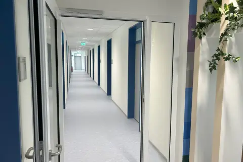 Langer Flur mit vielen Türen, beleuchtet von Deckenlampen. Grüne Notausgangsschilder sind sichtbar. Die Wartung von Feststellanlagen ist hier essenziell für die Sicherheit.