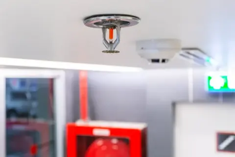 Eine Sprinkleranlage an der Decke, bereit für den Notfall. Daneben ein Rauchmelder und ein grünes Notausgangsschild.